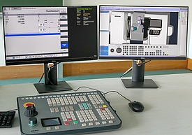Tisch mit zwei Bildschirmen, die verschiedene Ansichten der Simulationssoftware zeigen, davor liegen ein maschinentypisches Eingabetablet und eine Computermaus
