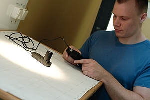 Ein Student schaut auf ein Messgerät, was er in der Hand hält. Der Messfühler ist unter einem Lichtkegel auf dem Versuchsstand platziert..