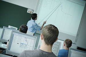 Das Bild zeigt den Blick in den Unterrichtsraum, auf die Bildschirme Richtung Tafel. Dort steht der Lehrende mit Zeigestock und erklärt.