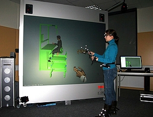 Eine Mitarbeiterin steht vor dem Virtual Reality System, in der Hand einen Flystick und Trackingpunkte am Körper, auf der Projektionswand ist ein Montagearbeitsplatz abgebildet